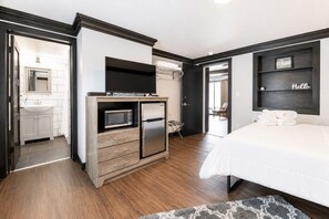 Habitació d'hotel moderna amb petita cuina, un llit i un bany visibles a través d'una porta oberta.
