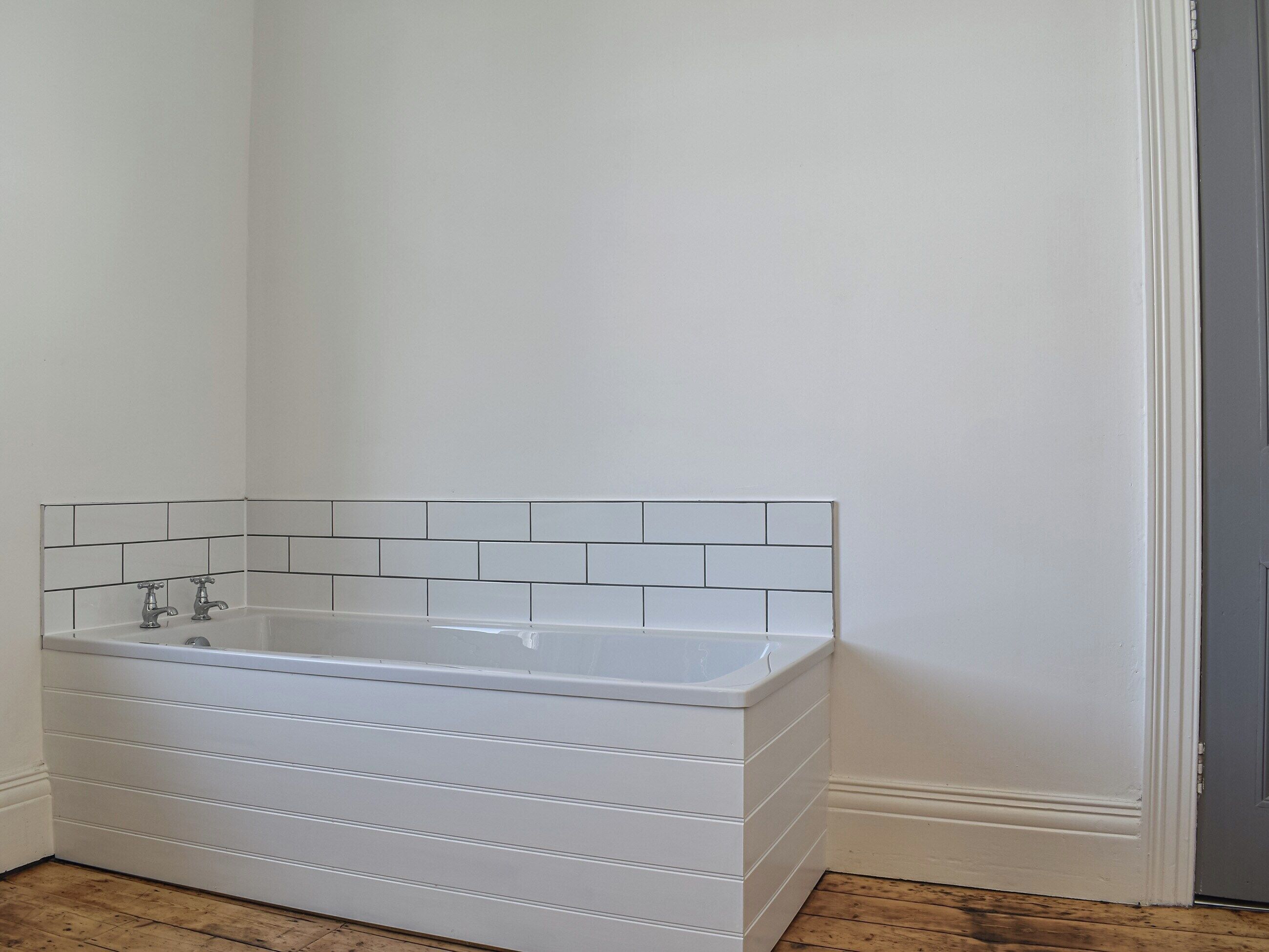Una bañera blanca con un revestimiento de azulejos en una esquina de una habitación con suelo de madera.