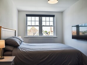 A bedroom with a bed, a nightstand, a lamp, a window with a view of buildings, and a TV mounted on the wall. - 8 bedroom accommodation in Swanage (Swanage)