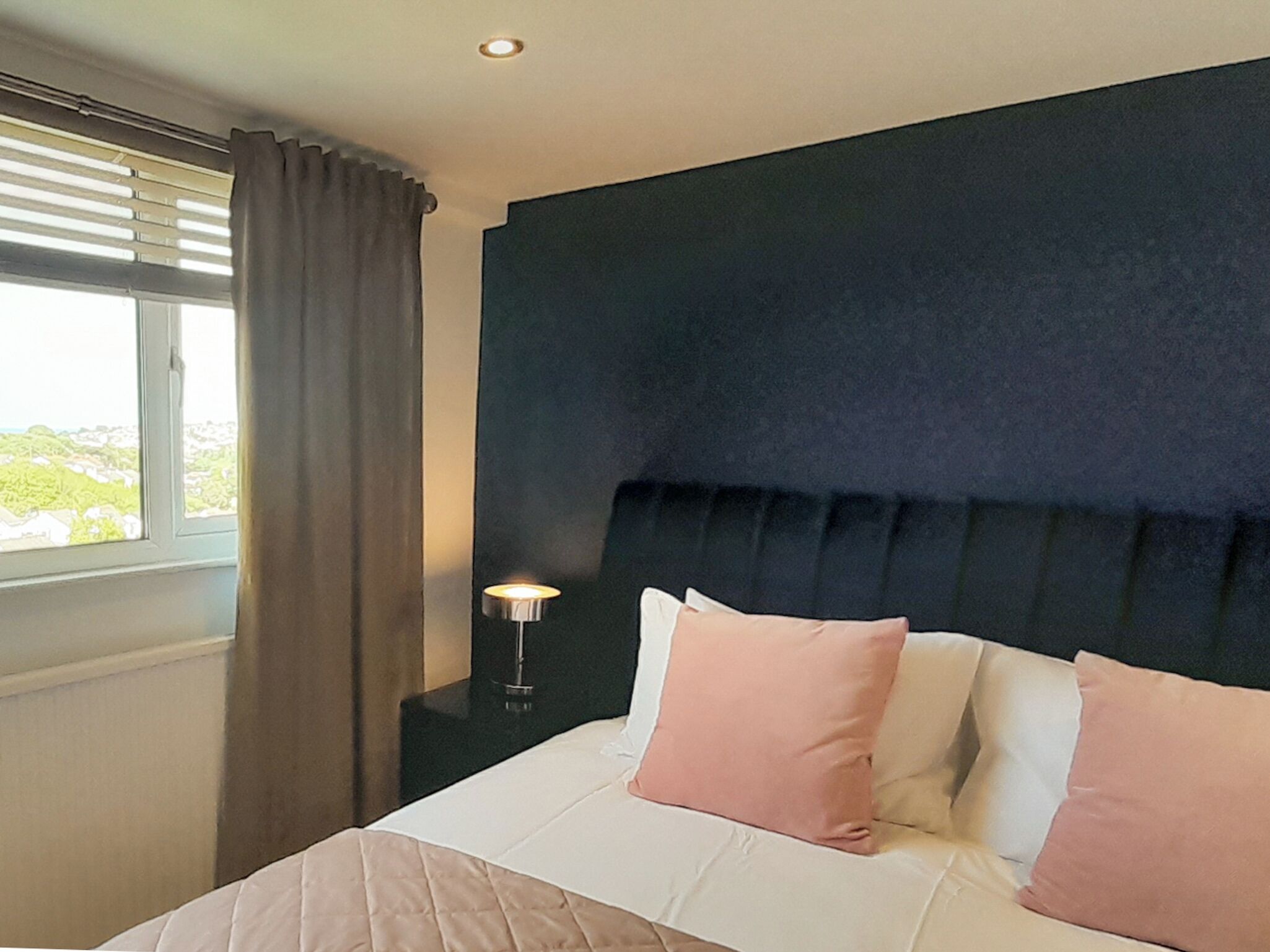 A neatly made bed with a quilted cover and two pillows, a bedside lamp, and a window with curtains.