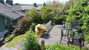 A garden patio with a table and chairs, surrounded by lush greenery and a white fence.