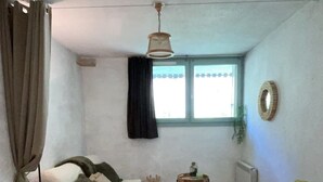Une petite pièce agréable, avec un plancher en bois, un canapé blanc recouvert d’une couverture à carreaux, une petite table avec une nappe verte et une fenêtre avec des rideaux.