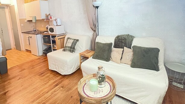 Un salon chaleureux avec un canapé, deux fauteuils, une petite table avec une bougie, et un coin cuisine à l’arrière-plan.
