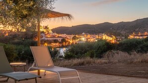 A wooden deck with two lounge chairs and a table, overlooking a hillside village at sunset.