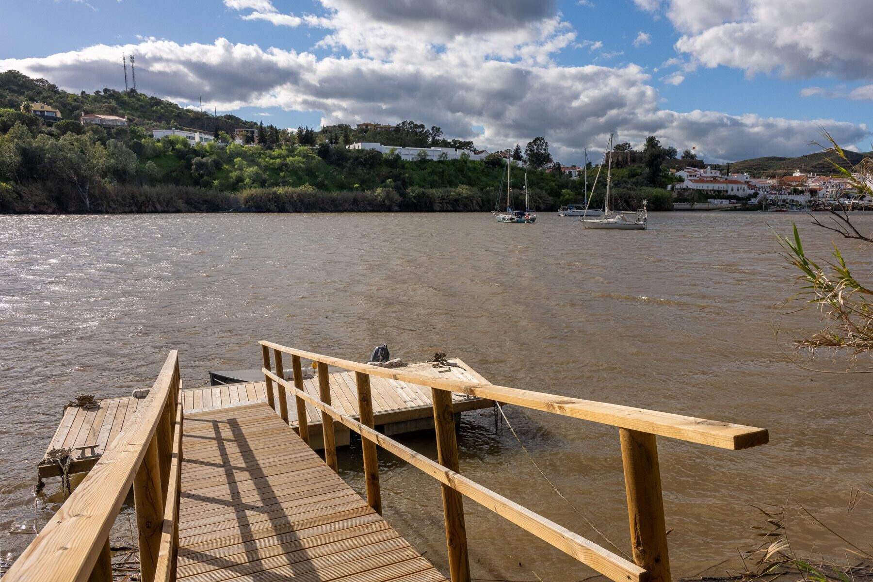 A wooden dock extending into a river with boats in the background and houses on the opposite bank.