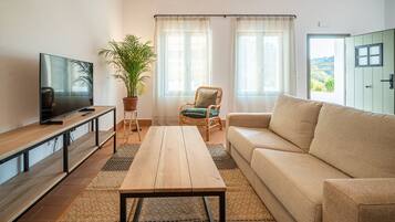 Uma sala de estar com televisão, mesa de centro de madeira, sofá, poltrona e um vaso com planta.