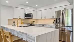 A modern kitchen with white cabinetry, a stainless steel refrigerator, a microwave, and a large island with bar stools. - Beautiful Fully Renovated Pool View Unit Just A Short Stroll To Beach Access! (Saint Augustine)