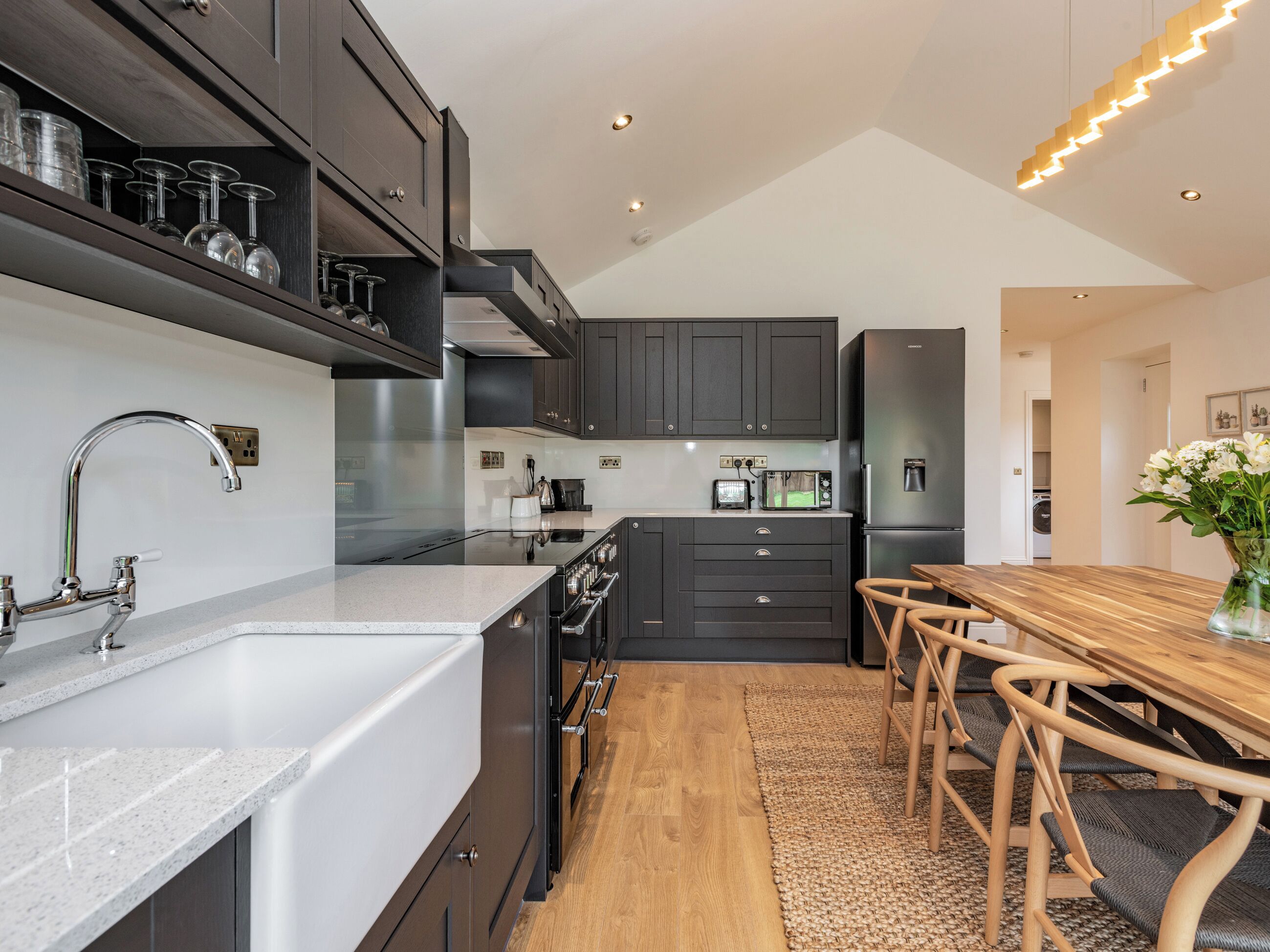 A modern kitchen with dark cabinets, a stainless steel range hood, a built-in oven, a coffee maker, and a dining area with a wooden table and chairs.