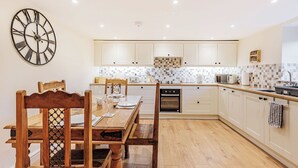 A modern kitchen with white cabinetry, a wooden dining table, and a wall-mounted clock. - 2 bedroom accommodation in Penybntfawr, near Oswestry (Penybntfawr, near Oswestry)