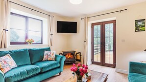 A living room with a teal sofa, a wooden coffee table with a vase of flowers, a fireplace, and a sliding glass door leading to an outdoor area. - 2 bedroom accommodation in Penybntfawr, near Oswestry (Penybntfawr, near Oswestry)