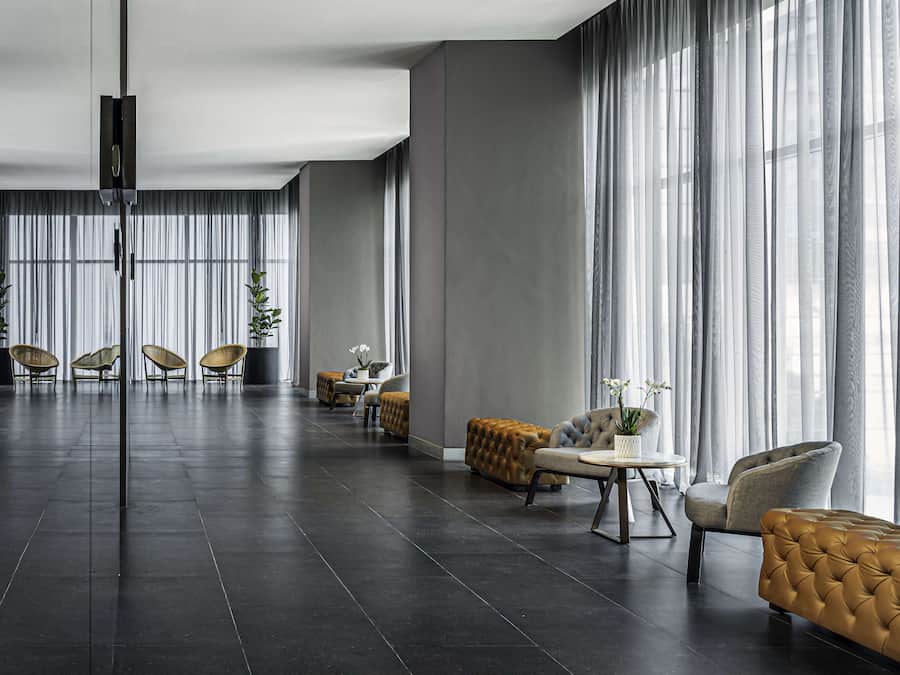 Una habitación amplia con piso de baldosas oscuras, ventanas grandes con cortinas translúcidas y una combinación de muebles modernos y clásicos, incluyendo sofás capitonados y sillas de mimbre.
