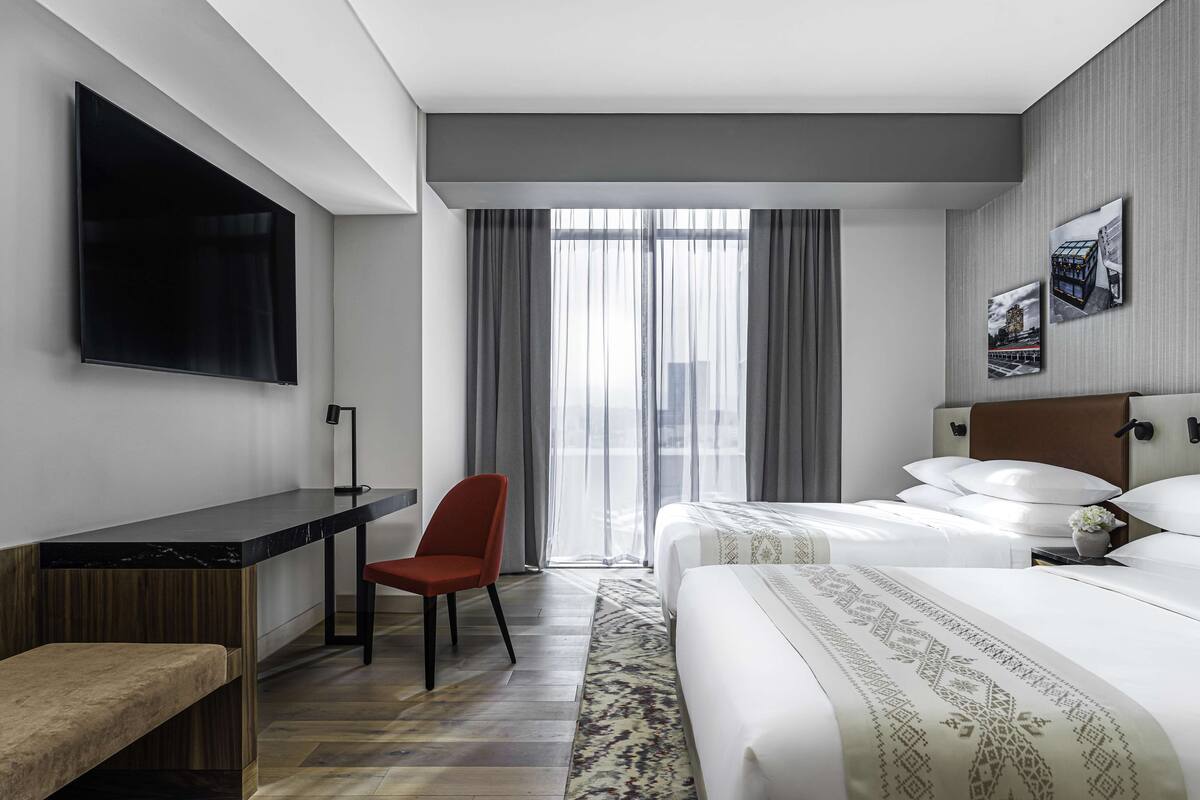A modern hotel room with a large bed, a desk with a chair, a flat-screen TV, and a large window with curtains.