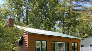 Una pequeña cabaña de madera con techo verde, rodeada de árboles y césped.