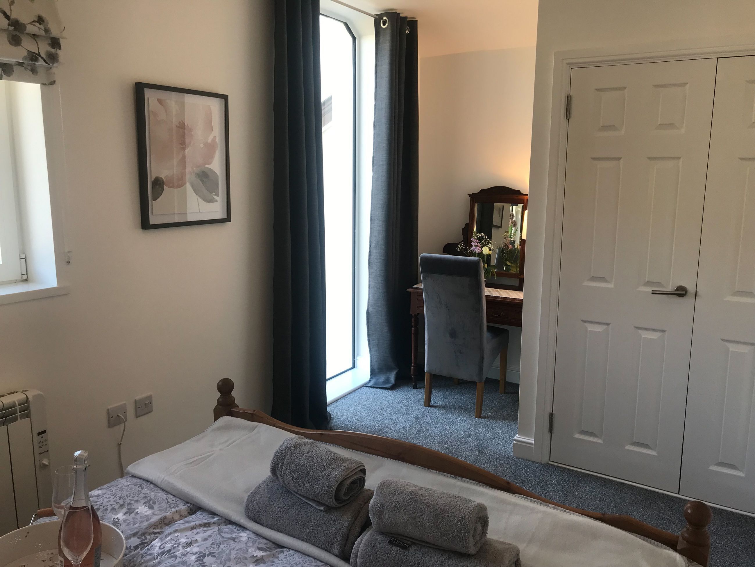 A hotel room with a bed, a desk with a chair, a window with curtains, a door, and a framed picture on the wall.