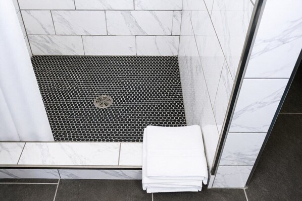 Zona de ducha con suelo de baldosas blancas, suelo de ducha hexagonal blanco y negro y una toalla blanca doblada en el suelo.