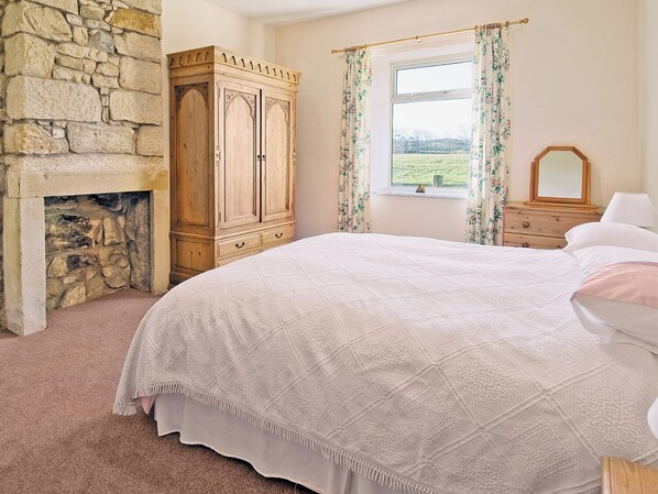 A bedroom with a stone fireplace, a large bed with a quilt, a wooden wardrobe, and a window with floral curtains.