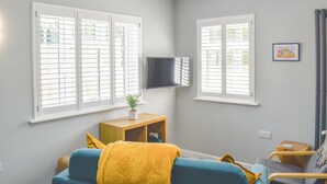 Ein Wohnzimmer mit zwei Fenstern, einem Flachbildfernseher, einer Couch, einem kleinen Holztisch und einem gerahmten Bild an der Wand.