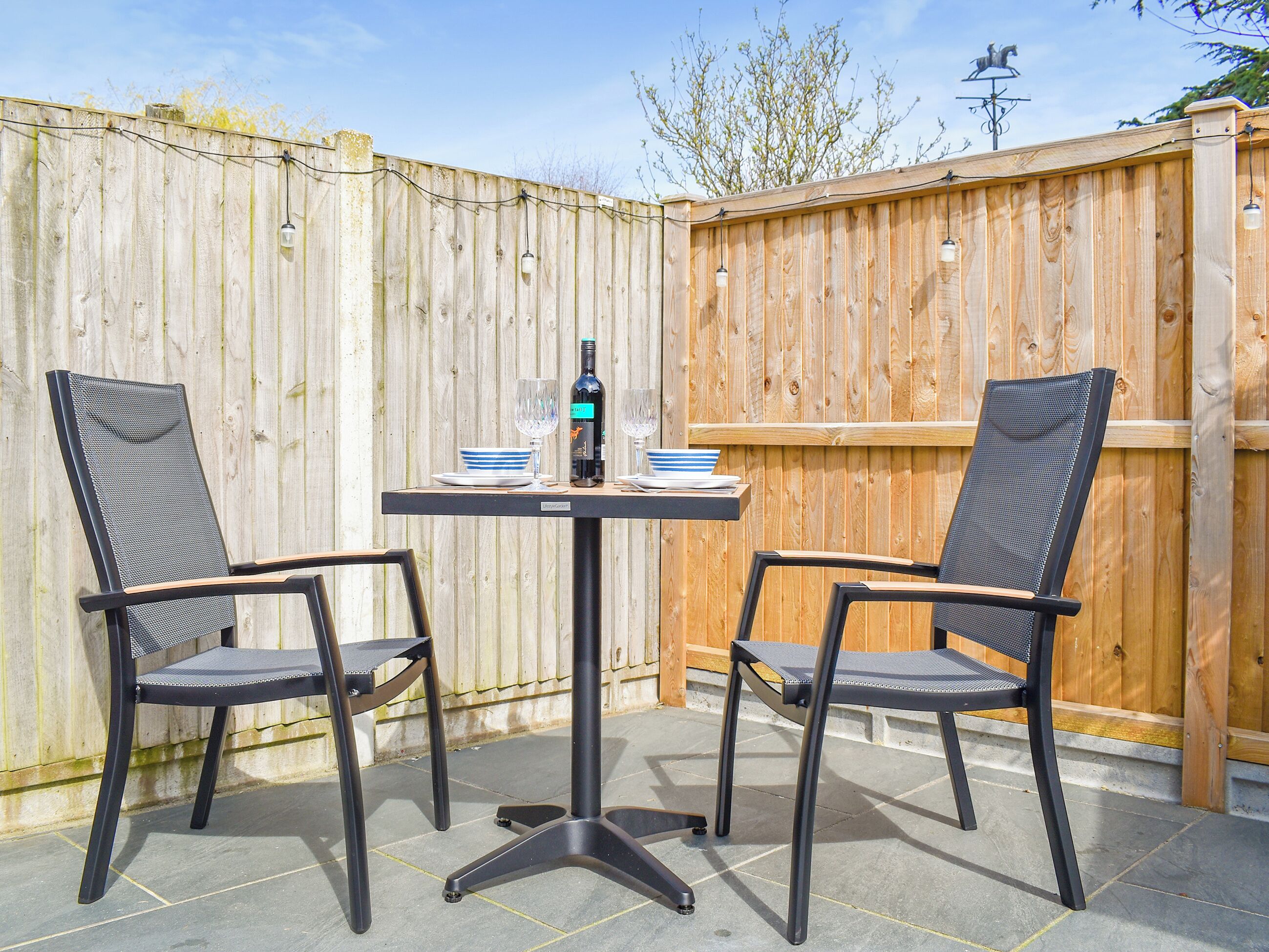 An outdoor patio with two chairs and a table, a bottle of wine, and a bowl of fruit.