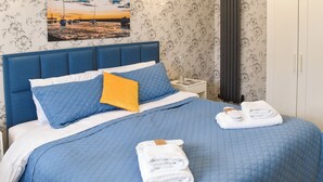 Ein ordentlich bezogenes Bett mit blauer Bettwäsche und einem gelben Kissen, ein weißer Nachttisch mit einer Lampe sowie ein gerahmtes Bild an der Wand.