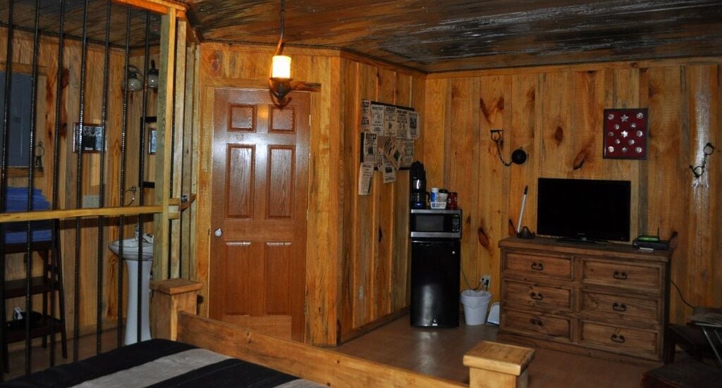 Interior de una cabaña de madera con puerta de madera, zona de cocina con microondas, cafetera y televisor sobre un mueble de madera.
