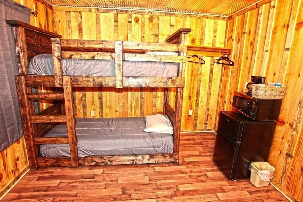 A bunk bed room with wooden walls and floors, a small black refrigerator, and a single hanging light.