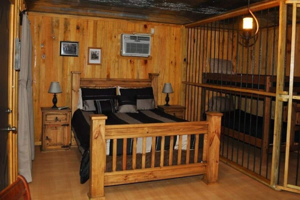 A wooden room with a bed, two bedside tables, a window, and a barred door.