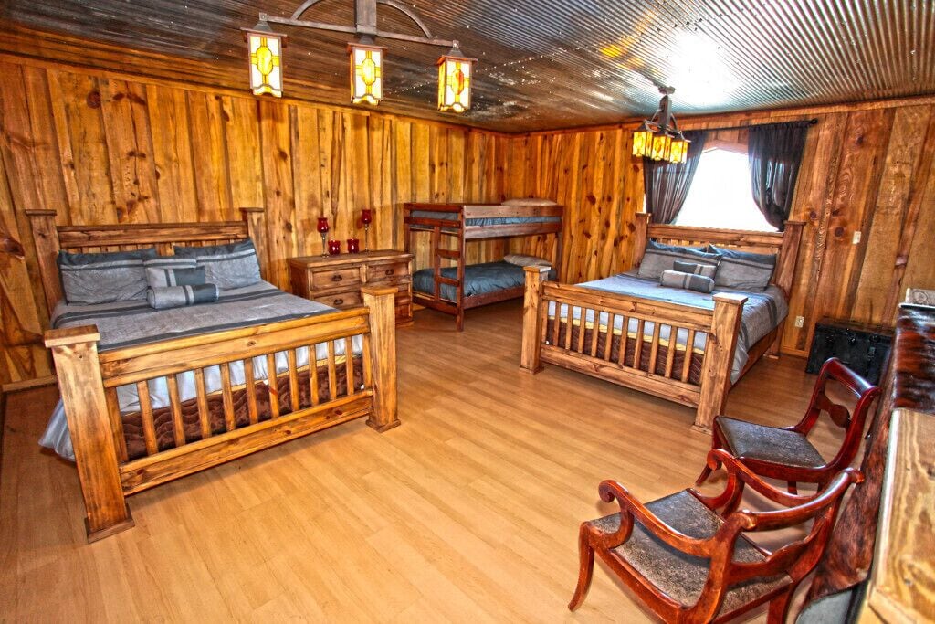 Una habitación rústica de madera con dos literas, un aparador de madera, una silla y una ventana con cortinas.