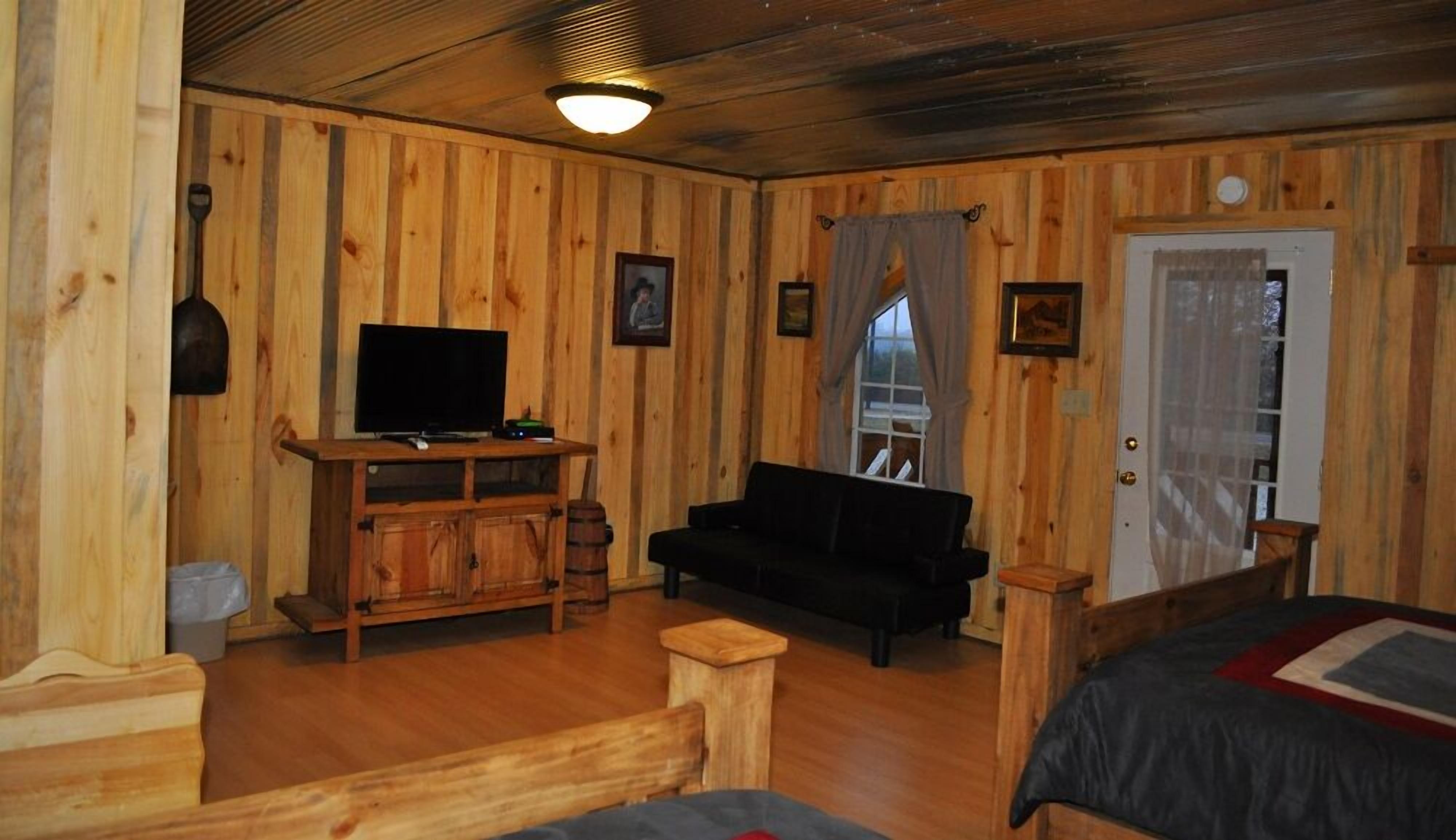L’intérieur d’un chalet en bois, avec un téléviseur posé sur un meuble en bois, un canapé noir et un lit recouvert d’une couverture rouge et bleue.