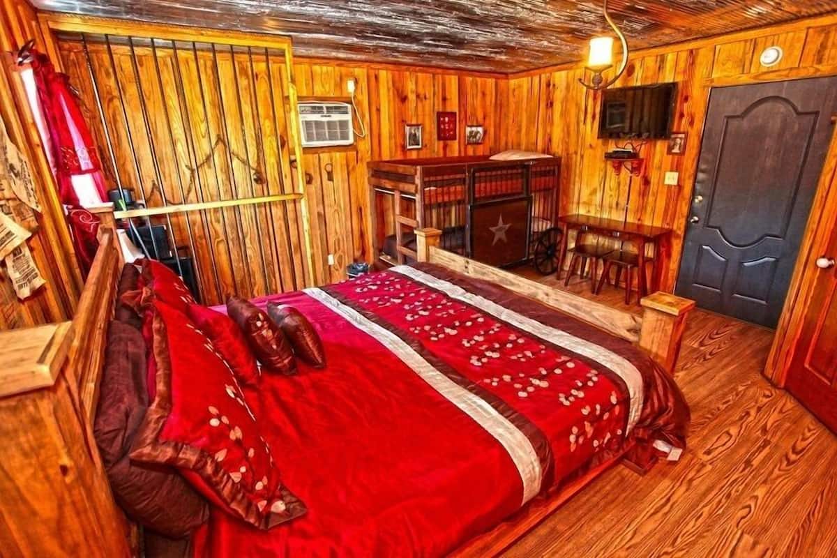 A wooden room with a bed, a small table, a chair, a fireplace, and a door.