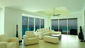 A spacious living room with a large L-shaped sofa, a coffee table, and a ceiling fan. Large windows provide a view of the ocean.