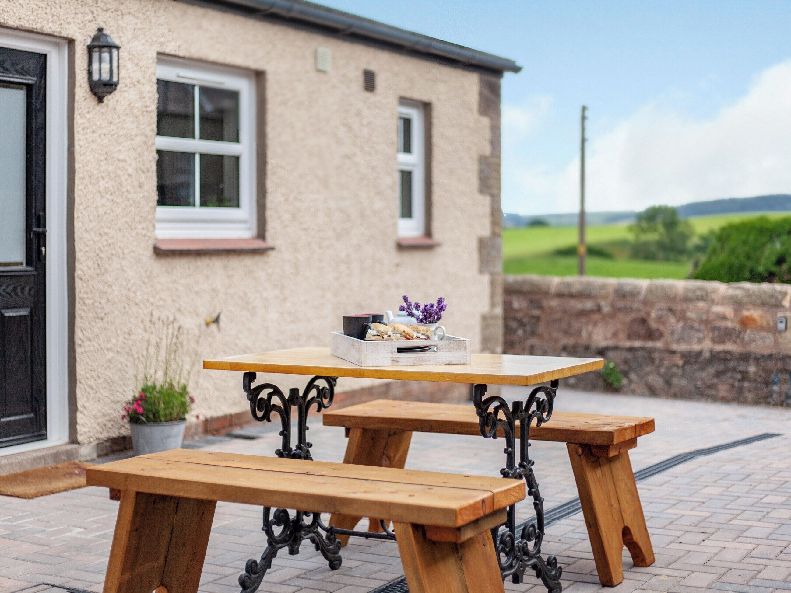 A wooden picnic table with benches outside a house with a stone wall and a view of the countryside.