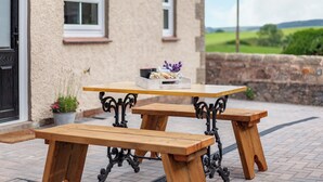 Une table de pique-nique en bois, avec des bancs, se trouve à l’extérieur d’une maison dotée d’un mur en pierre et offrant une vue sur la campagne.