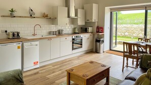 Une cuisine moderne avec des armoires blanches, un four encastré et un four à micro-ondes. On y trouve un coin repas avec une table et des chaises en bois, et une vue sur un paysage verdoyant grâce à de grandes fenêtres.