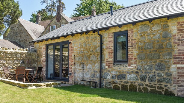 Un bâtiment en pierre avec un toit en tuiles, une porte vitrée et un petit espace extérieur aménagé avec des chaises.