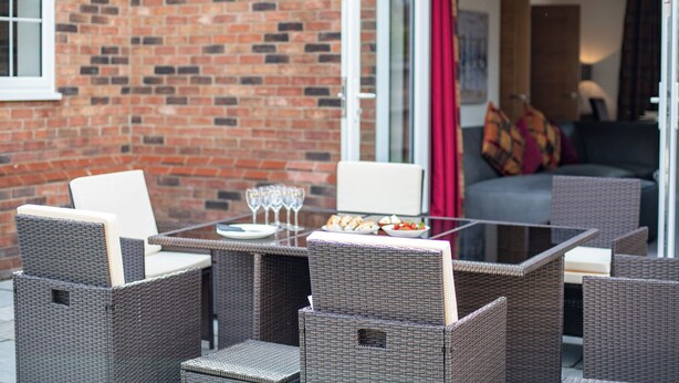 A patio with wicker furniture, a glass table, and a brick wall.