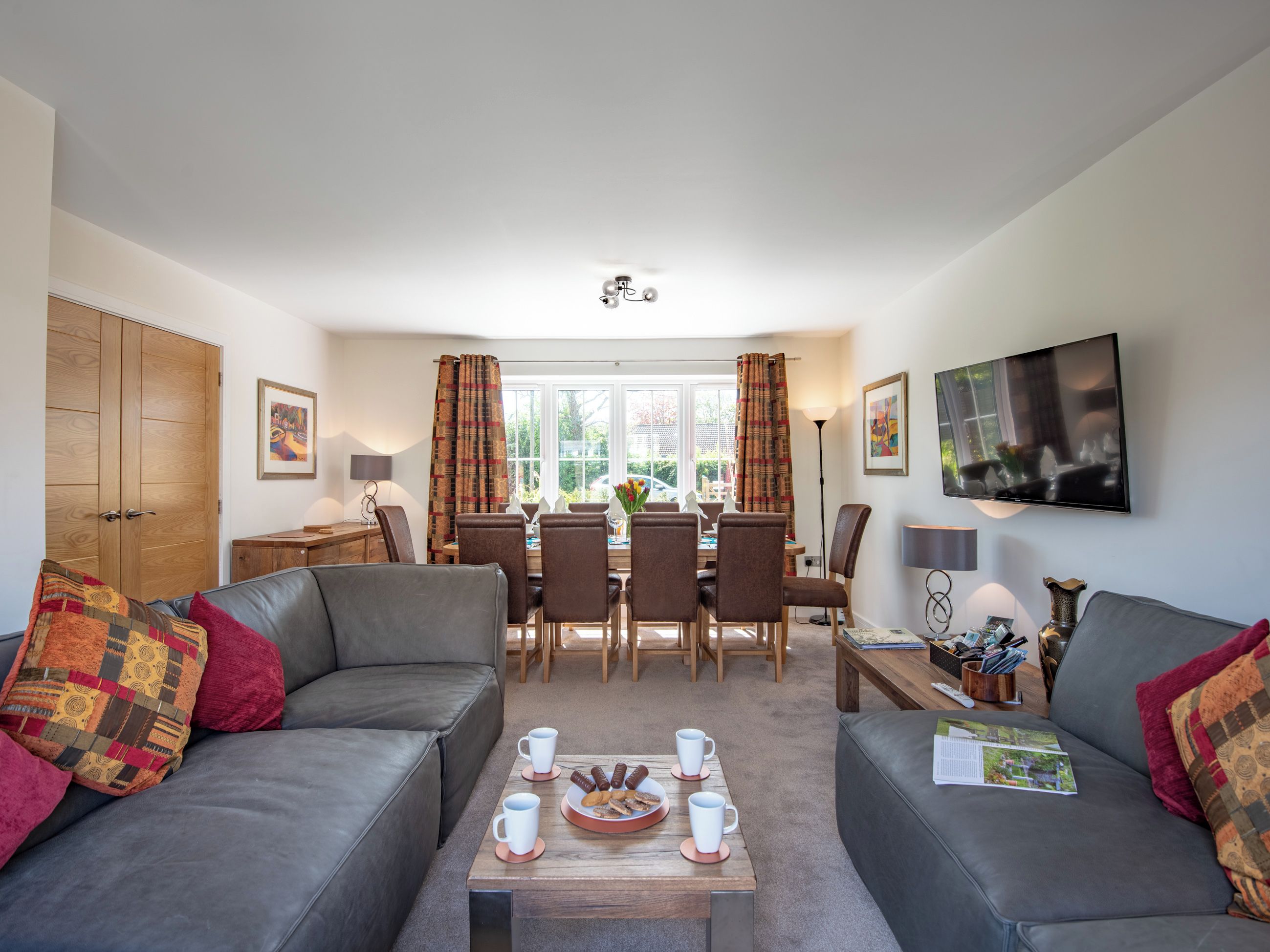 Ein Wohnzimmer mit einer Couch, einem Couchtisch mit Gebäck, einem Esstisch mit Stühlen und einem an der Wand befestigten Fernseher.