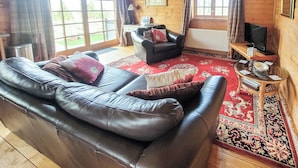 A cozy living room with a leather sofa, a black armchair, and a red patterned rug. There is a wooden TV stand with a television and a small table with a bowl and papers. - 2 bedroom accommodation in Kippford near Dalbeattie (Kippford near Dalbeattie)