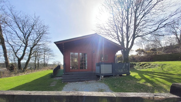 A small red cabin with a large window, surrounded by a grassy area and trees. - 2 bedroom accommodation in Kippford near Dalbeattie (Kippford near Dalbeattie)