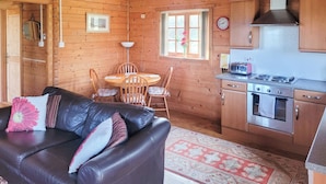 A cozy cabin interior with a wooden dining area, a leather sofa, a kitchen with a stove and microwave, and a large window with curtains. - 2 bedroom accommodation in Kippford near Dalbeattie (Kippford near Dalbeattie)