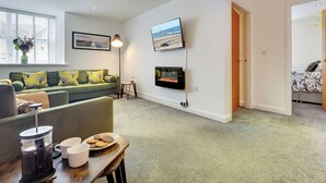A modern living room with a green sofa, a coffee table with a coffee maker and cookies, a TV mounted on the wall, and a window with blinds. - 2 bedroom accommodation in Berwick-upon-Tweed (Berwick-upon-Tweed)