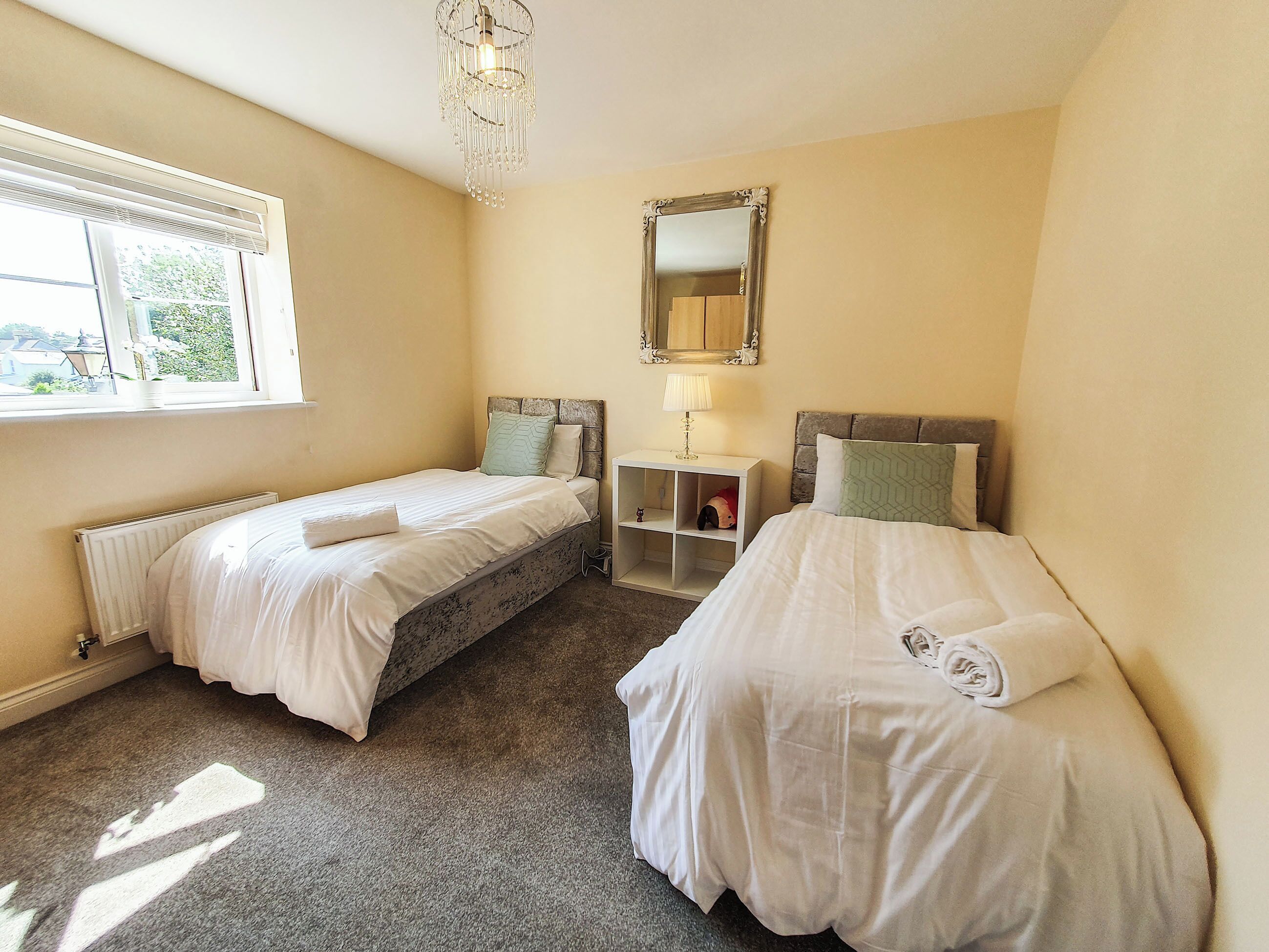 A hotel room with two beds, a window with blinds, a radiator, a bedside table with a lamp, and a mirror on the wall.