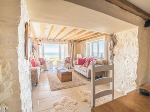 Habitació acollidora amb paret de pedra, bigues de fusta al sostre, seients de sofà, un arca de fusta i vistes al mar.