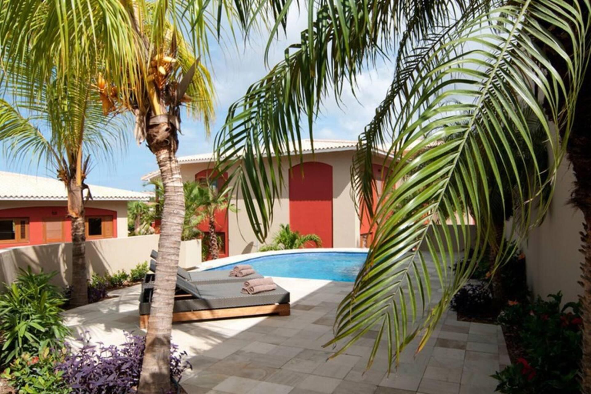 A pool area with a lounge chair, palm trees, and a building with red and white walls.