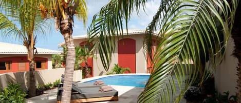 A pool area with a lounge chair, palm trees, and a building with red and white walls.