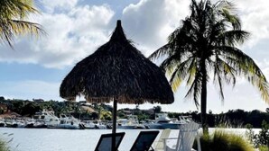 A beach with a thatched umbrella, two lounge chairs, and a palm tree. Boats are visible in the background. - Appartement La Maya met Zwembad Strand Jan Thiel (Jan Thiel)