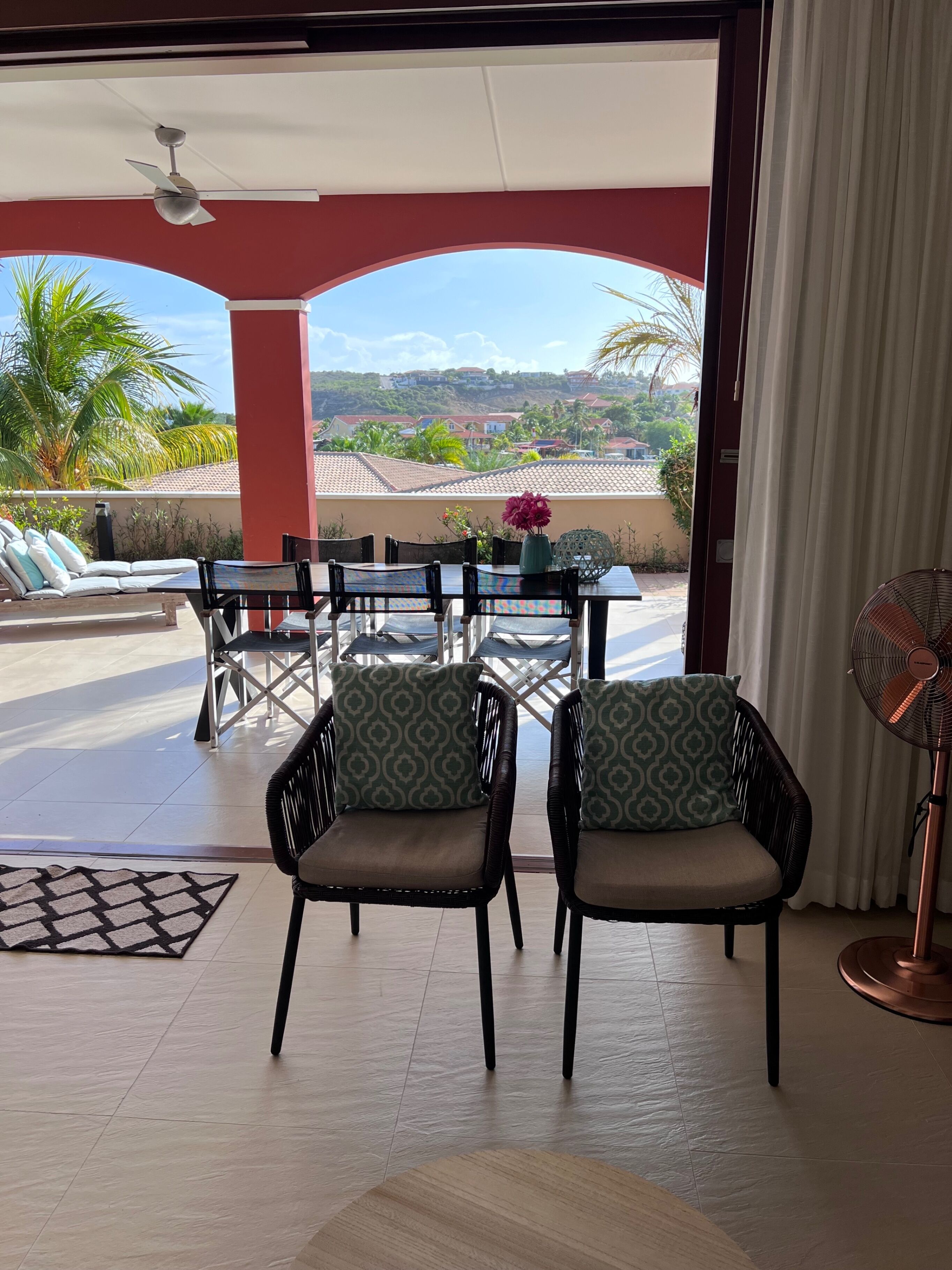 Un patio luminoso con tavolo e sedie, una ventola a soffitto e vista su una zona residenziale con palme e case.