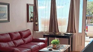 A room with a red sofa, a small table with a plant, a TV, and large windows with curtains. - Katie's Cozy Suites (Tombstone)