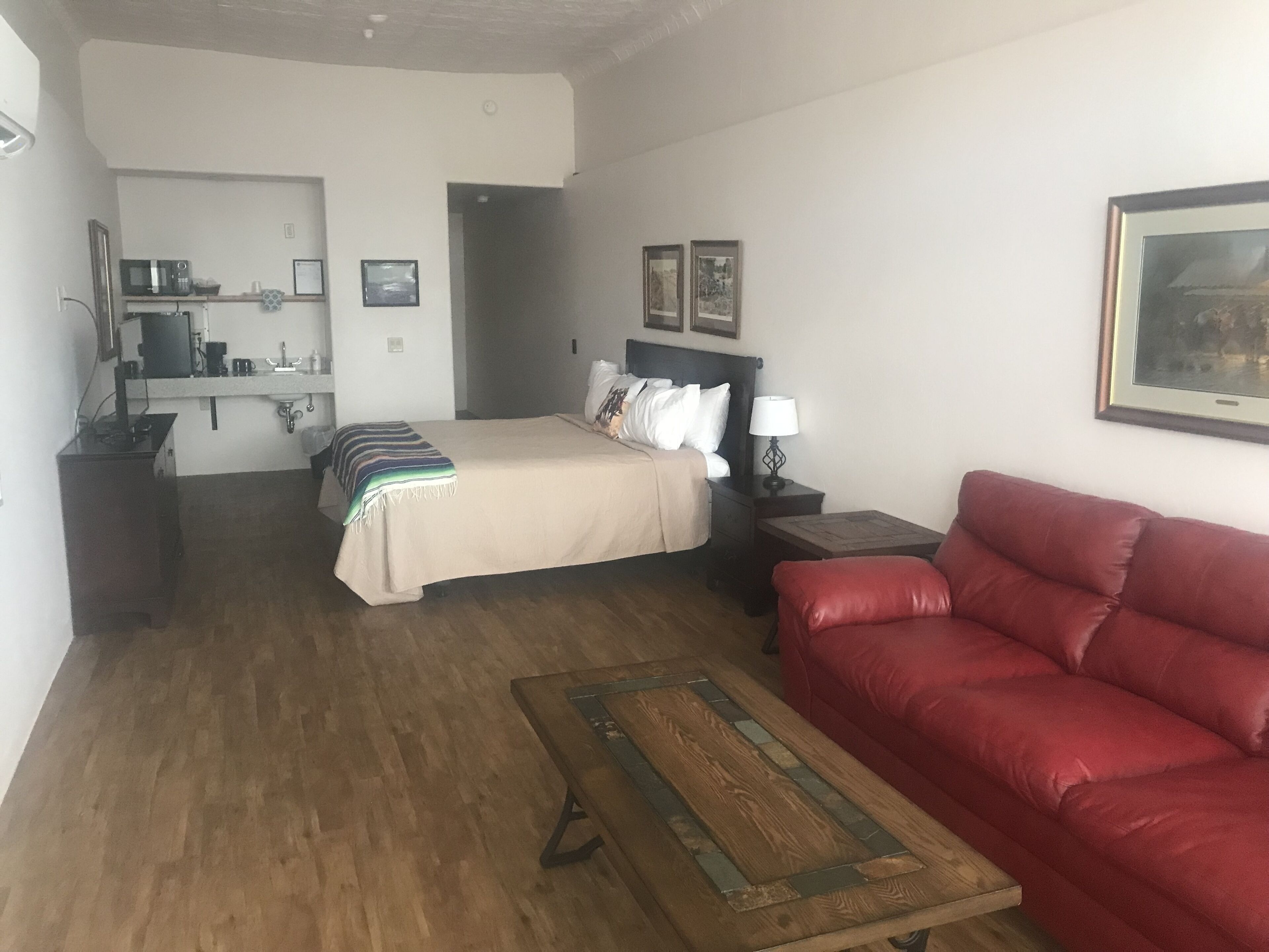 Habitación de hotel con cama, sofá rojo, mesa de centro de madera, mesita de noche con lámpara, pequeña mesa con microondas y estante con microondas y otros objetos.