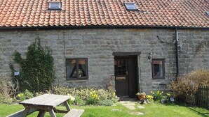 A stone building with a tiled roof, a wooden door, and a small garden with a picnic table. - Nestled in a prime location with spectacular views over open countryside. (Staintondale near Scarborough)