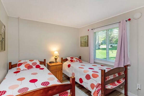 A bedroom with two single beds, a nightstand with a lamp, a window with curtains, and a painting on the wall. - Dog Friendly Smugglers` Notch Relaxing Mountainside Retreat (Cambridge)